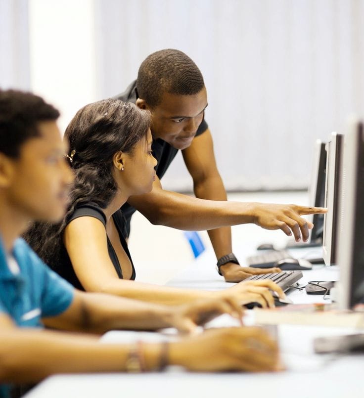 Learners collaborating at computers in a classroom, with a tutor pointing at a screen
